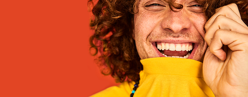 A man with curly hair wears a yellow turtleneck and smiles