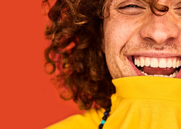 A man with curly hair wears a yellow turtleneck and smiles